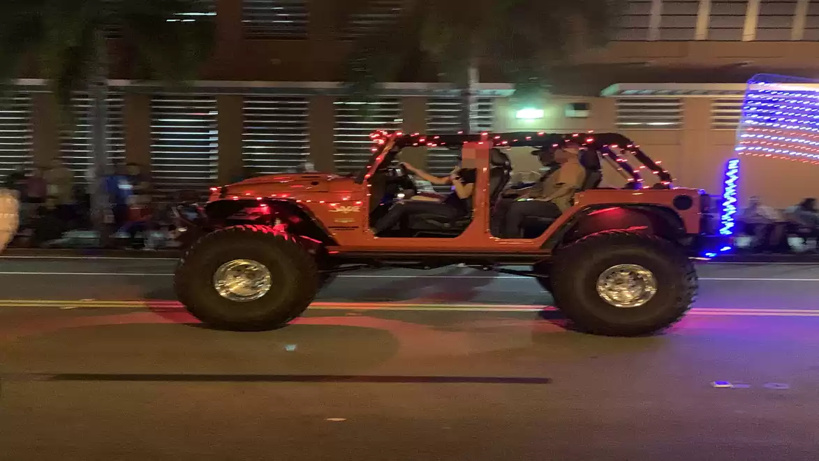A red Jeep, decorated with lights, drives on a street at night. Buildings and blurred people are visible in the background.