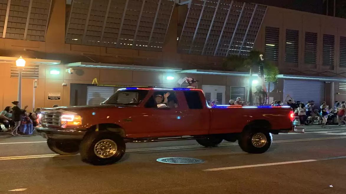 A red pickup truck driving on a street at night, surrounded by spectators.