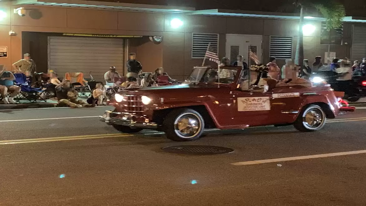 A red vintage convertible car drives along the street during a nighttime parade, with spectators on the sidewalk.