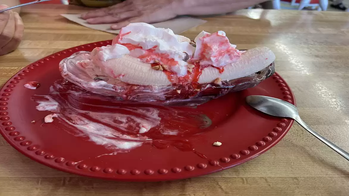 A banana split with whipped cream and strawberry sauce on a red plate, with a spoon beside it.
