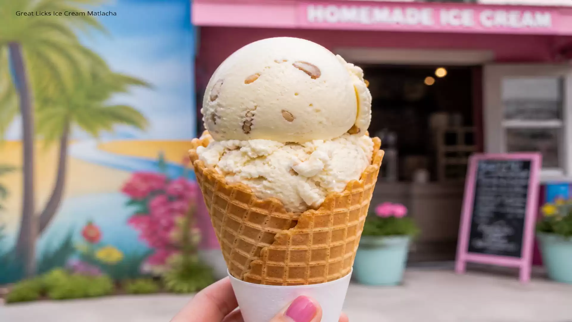 A hand holding a cone with two scoops of ice cream in front of an ice cream shop with colorful mural in the background.