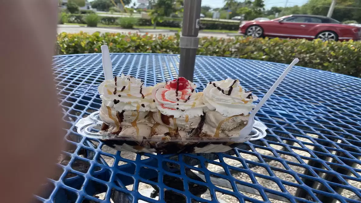 A banana split on a blue outdoor table, topped with whipped cream, chocolate sauce, and a cherry.