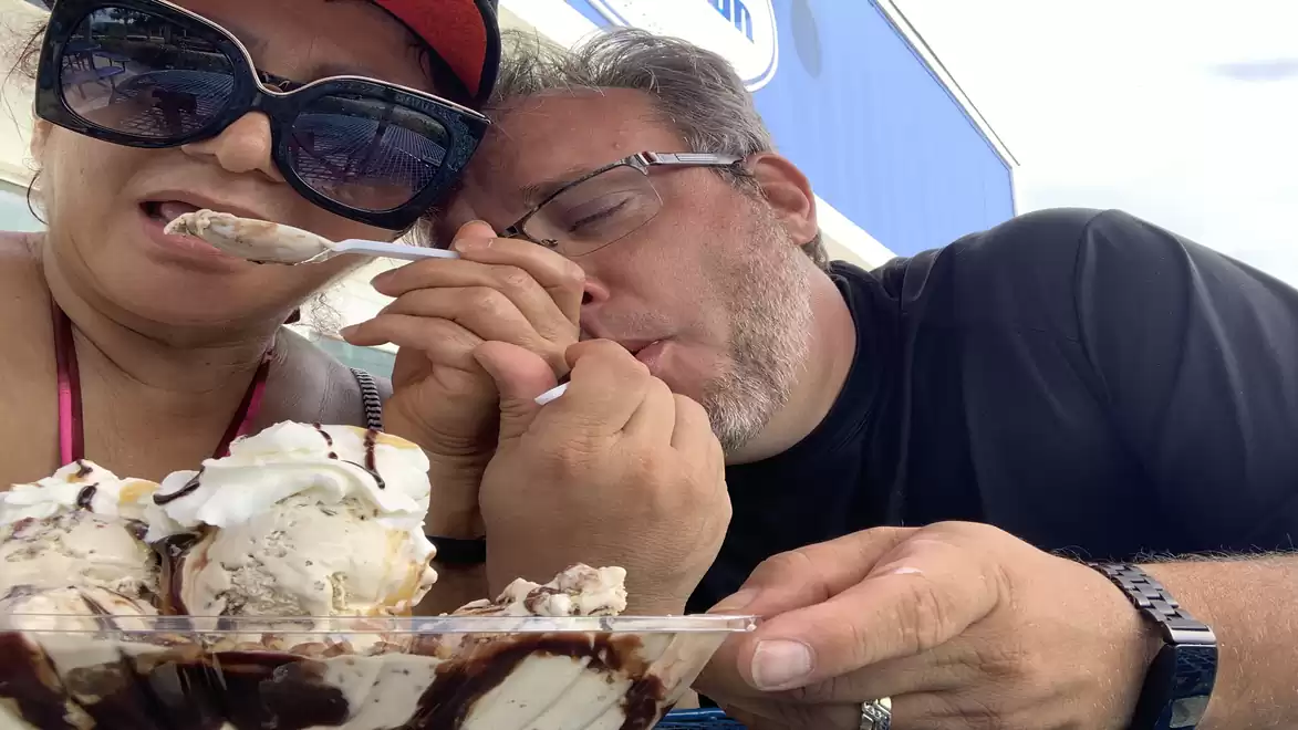 A man and a woman sit outside sharing a large ice cream with chocolate sauce.