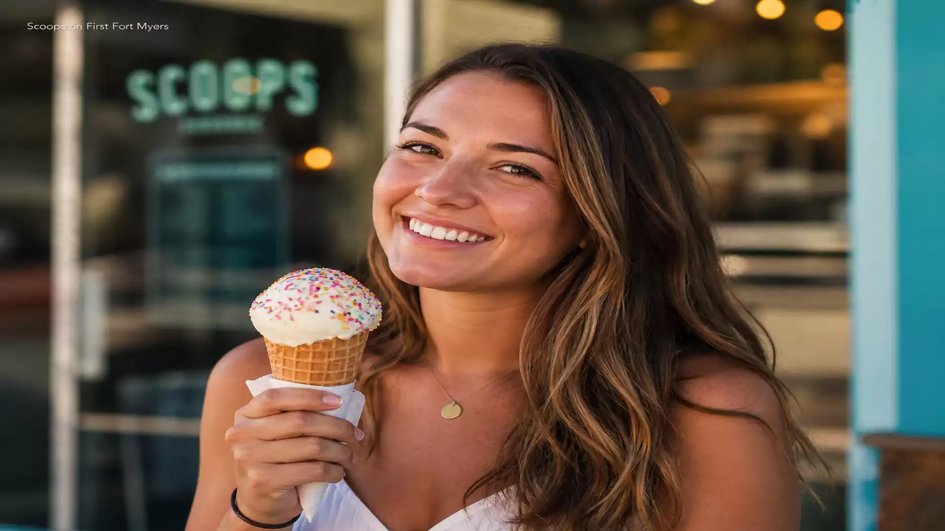 Woman smiling while holding an ice cream cone, with a shop in the background.