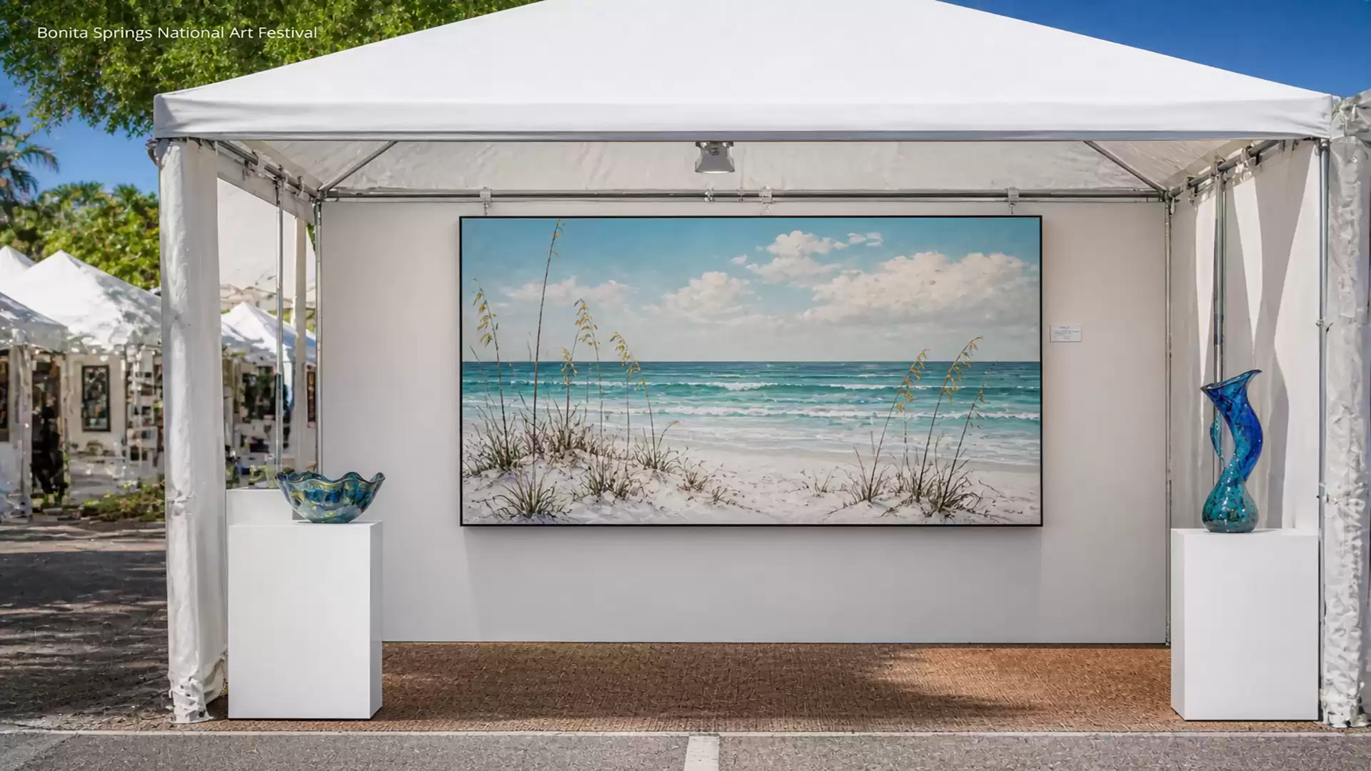 An art tent at a fair displays a large painting of a beach with a blue sky and white clouds. Glass decorations are placed on both sides of the painting.