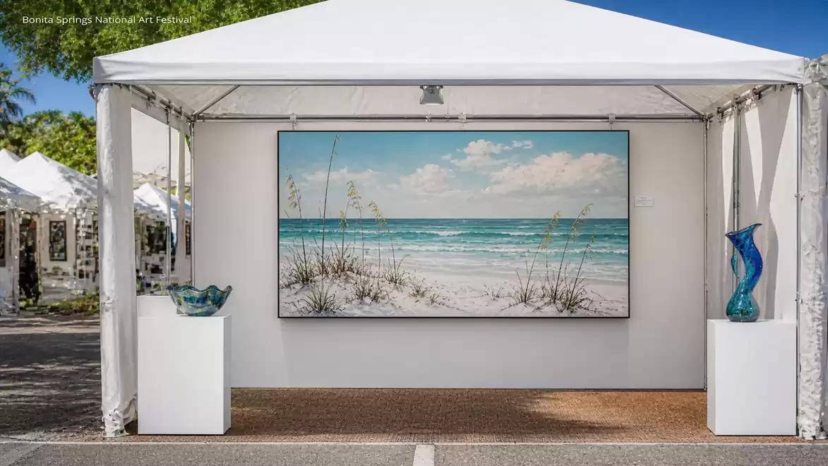 An art tent at a fair displays a large painting of a beach with a blue sky and white clouds. Glass decorations are placed on both sides of the painting.