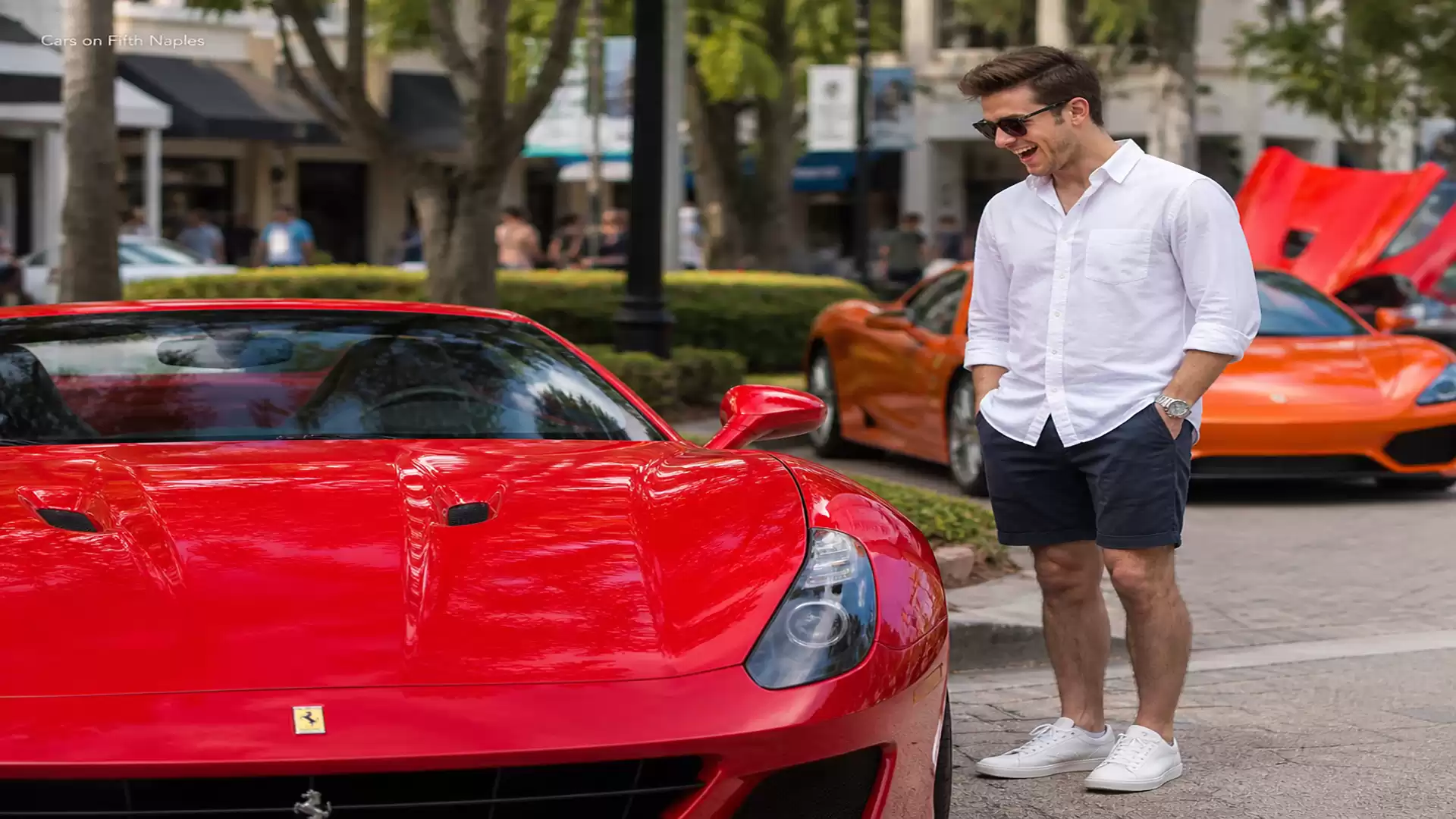 A man in a white shirt and shorts stands in front of a red sports car on a city street. In the background, more luxury vehicles are visible.