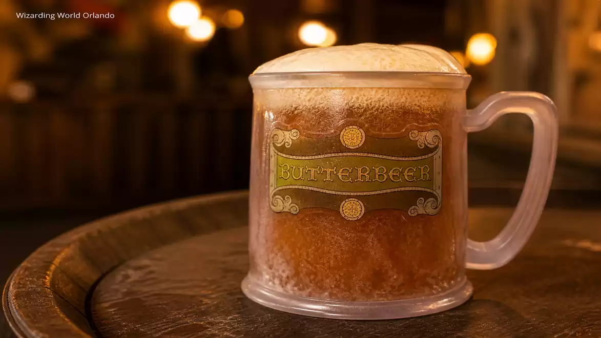 A mug of Butterbeer, filled with a frothy top, sits on a rustic wooden table in a cozy, warmly lit setting.