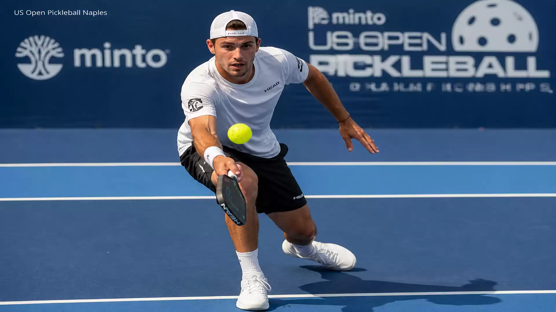 A male player in sports attire focused on a yellow ball during a pickleball game on a blue outdoor court.