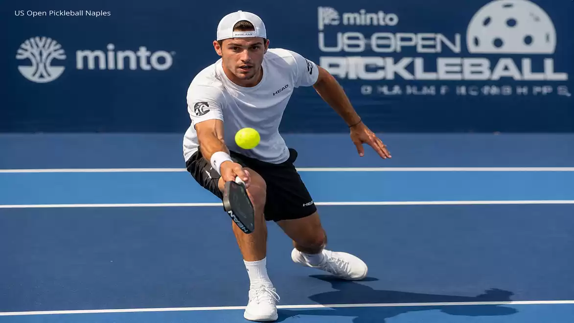 A male player in sports attire focused on a yellow ball during a pickleball game on a blue outdoor court.