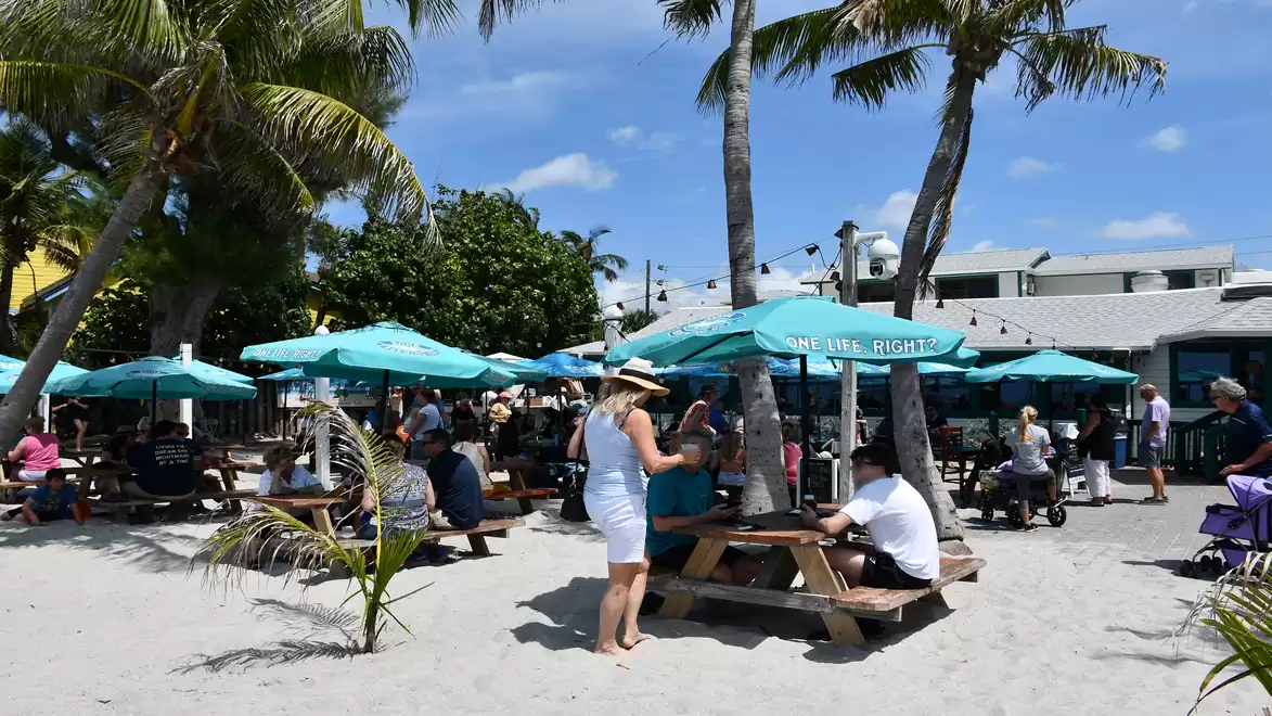 Beach scene with people sitting at picnic tables under palm trees and sun umbrellas. Blue sky with scattered clouds in the background.