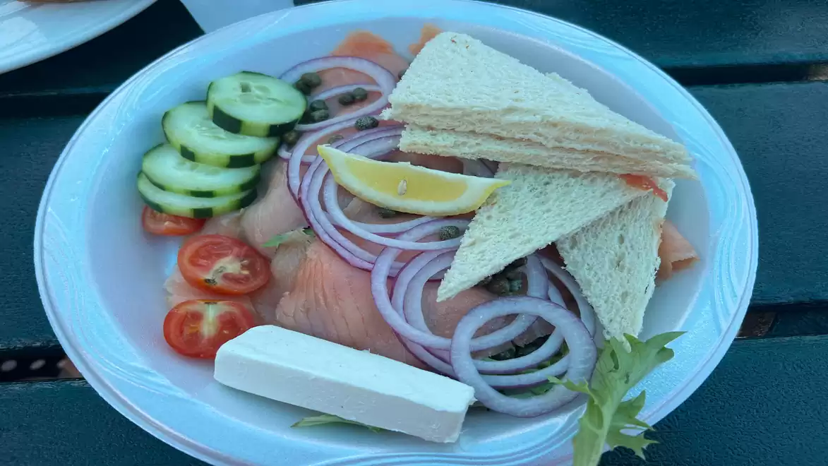 A plate with a selection of fresh ingredients, including cucumber slices, cherry tomatoes, onion rings, smoked salmon, a lemon wedge, toast triangles, and a piece of cream cheese, served on a dark wooden table.