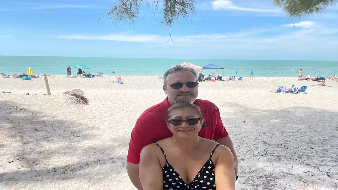 A couple posing for a selfie at the beach with bright sand and blue sky in the background. Other beachgoers and the sea are visible in the background.