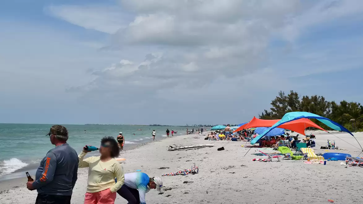 A long sandy beach with blue sky and clouds, where people relax under colorful sunshades.