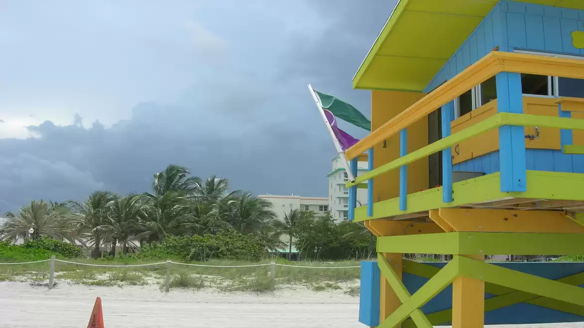 Colorful lifeguard hut on the beach under a cloudy sky, with palm trees in the background.
