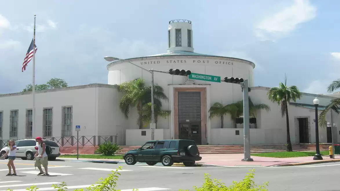 Historic post office building with Art Deco architecture on a street corner, lined with palm trees, with passing cars and pedestrians.