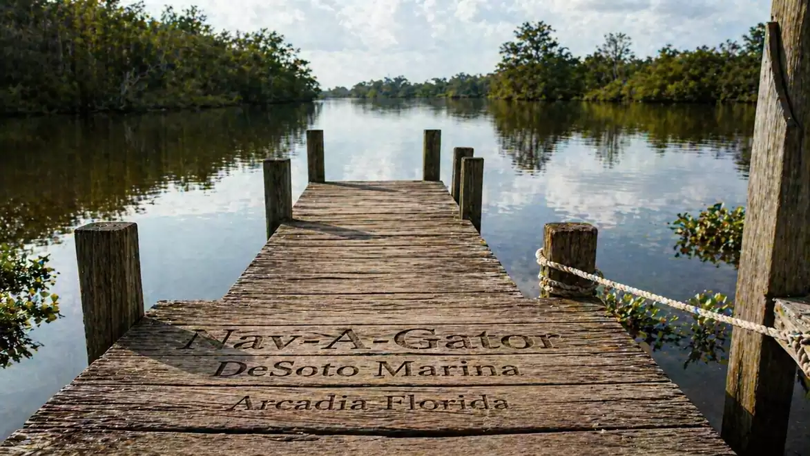 Wooden dock on a calm river with trees on both banks and a cloudy sky, Nav-A-Gator DeSoto Marina in Arcadia, Florida.