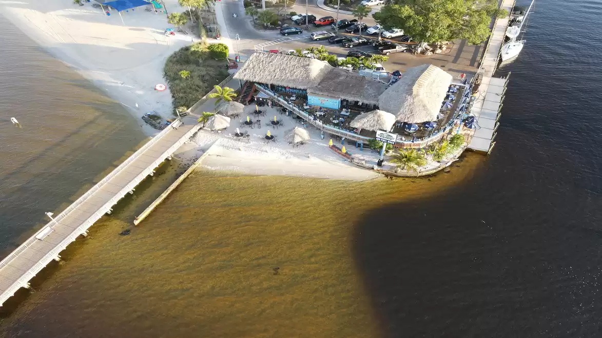 Aerial view of a beach with a pier, a building with a thatched roof, and several umbrellas near the water.