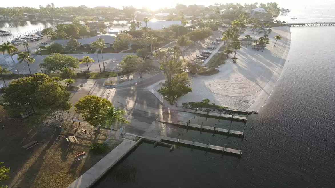 Aerial view of a coastal area with a beach, dock, and numerous trees, as the sun shines in the background.