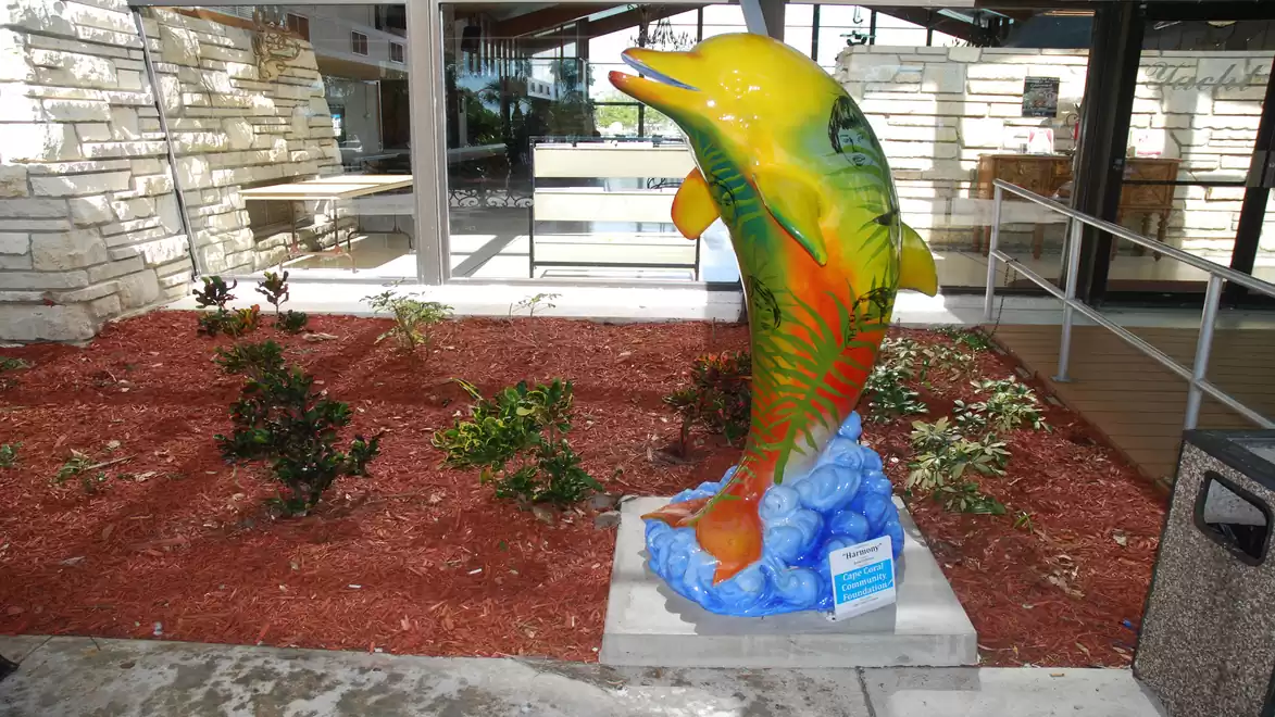 Colorfully painted dolphin statue in an outdoor area near a building with plants in the background.