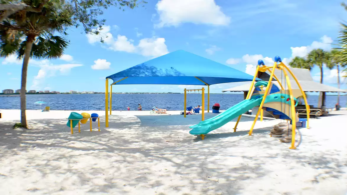 A sunny beach with a children's playground featuring slides and climbing structures. The white sand and blue sunshades create an inviting atmosphere right by the water.