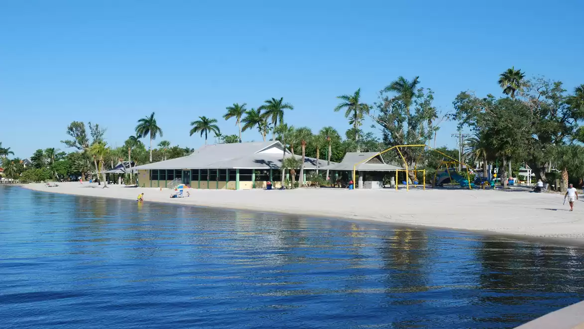 A sunny beach with palm trees and a building in the background, surrounded by blue sky and calm water.