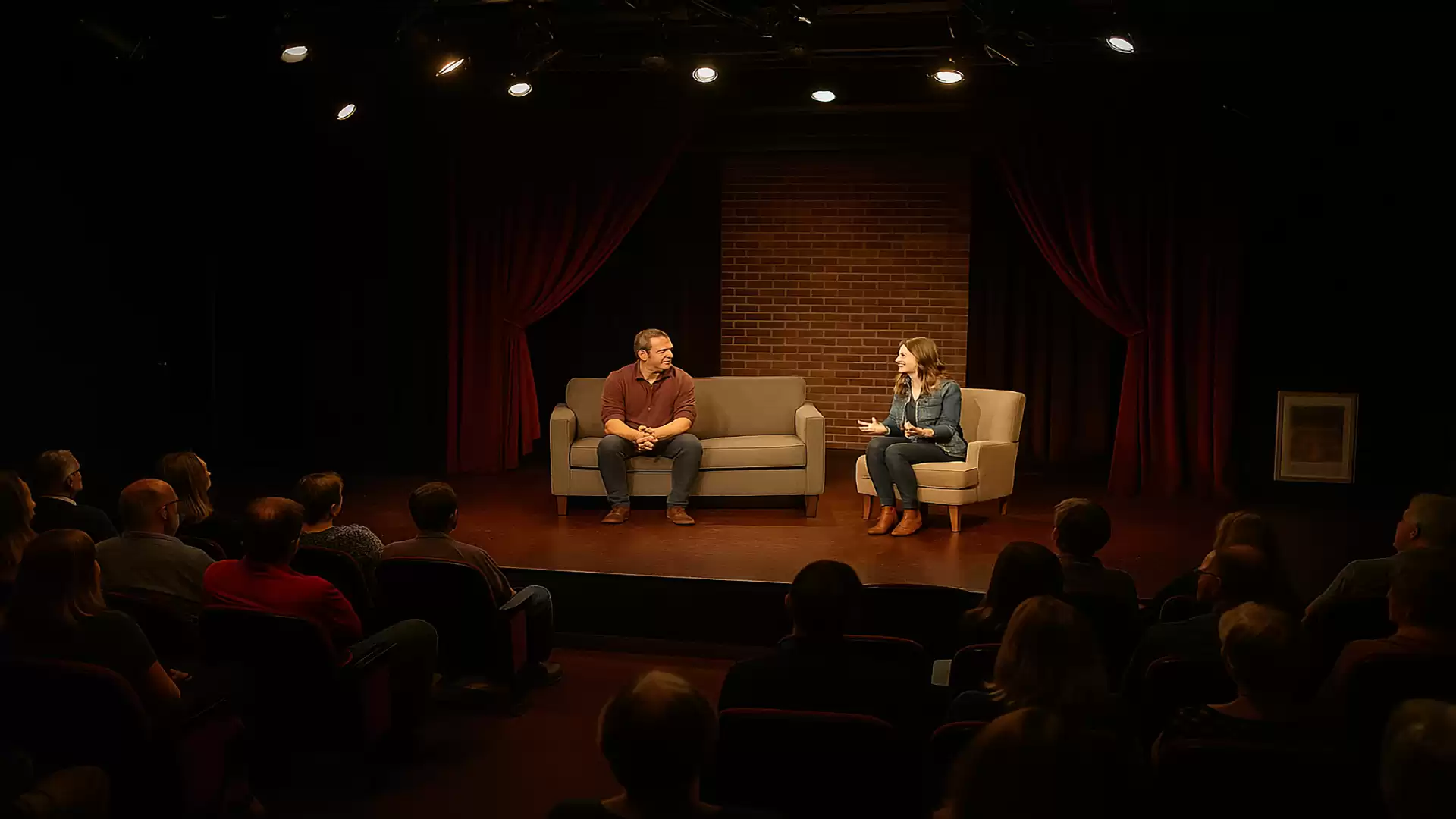 Two people sitting on a stage in front of an audience. One person is on a sofa, the other on a chair. The stage has a red curtain and a brick wall in the background.