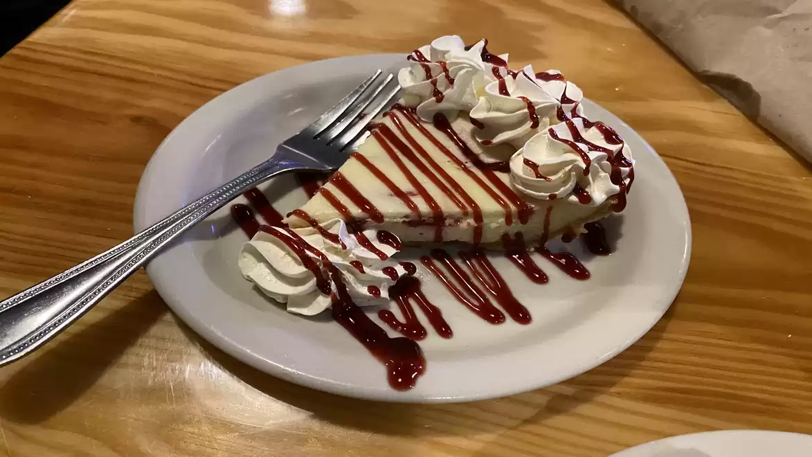 A slice of cheesecake on a white plate, garnished with whipped cream and raspberry sauce on a wooden table.