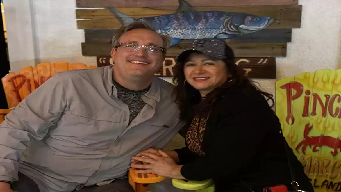 A man and a woman sitting together laughing on colorful chairs in front of a fish-themed sign in a restaurant.