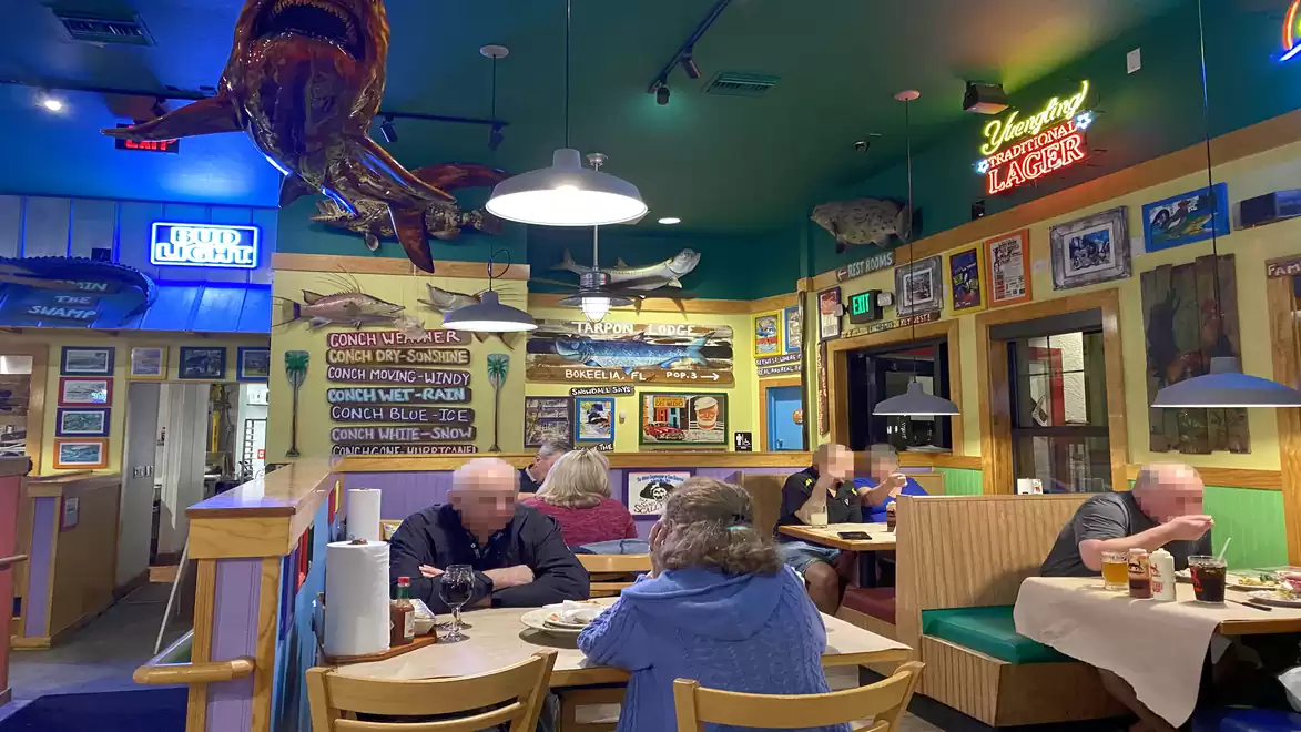 Interior view of a brightly decorated restaurant with customers at tables, fish decorations, and neon signs on the walls.