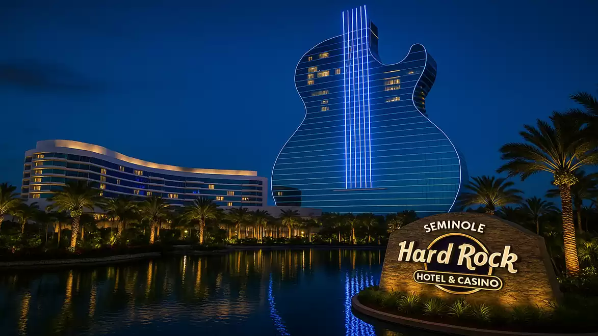 The image shows the Seminole Hard Rock Hotel & Casino at night. The building is shaped like a guitar and illuminated with blue lights, next to a lake and palm trees.