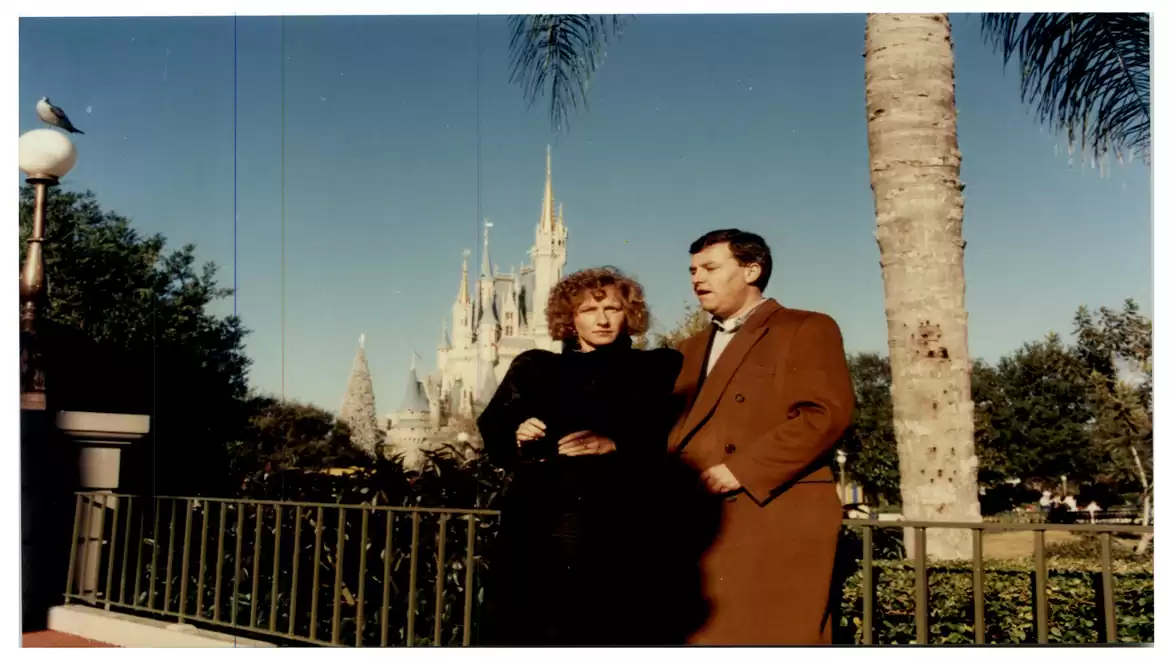 A man and a woman stand in front of a castle in an amusement park on a sunny day.