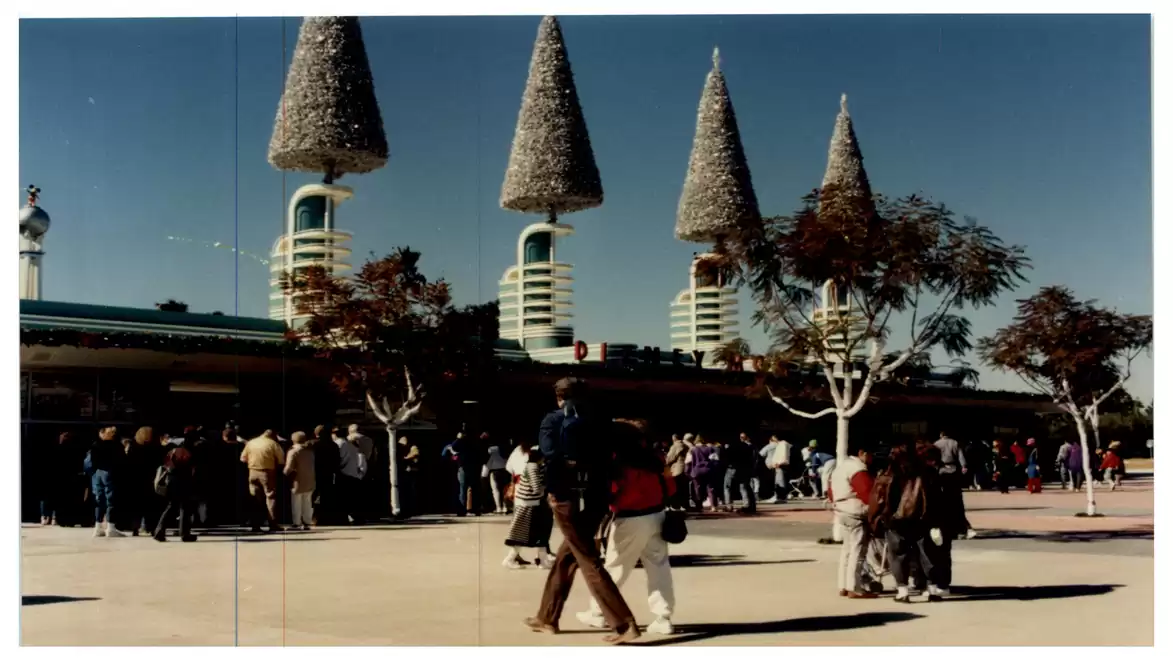A crowd stands under a blue sky in front of futuristic structures with pointed towers and trees.