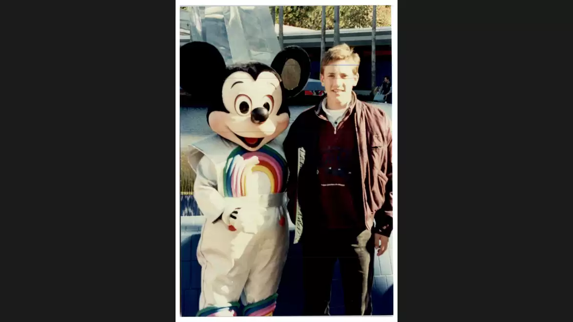 Person stands next to a large plush costume of Mickey Mouse outdoors in daylight.