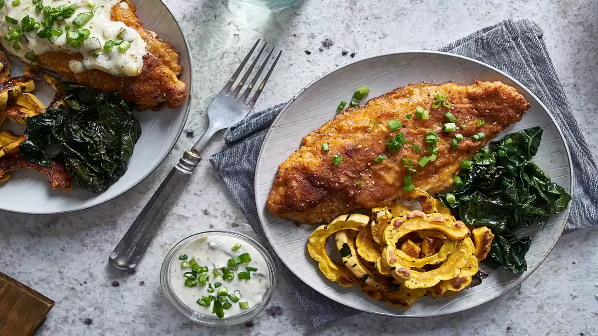 Two plates with breaded fish, sautéed leafy greens, pale squash, and herb sauce on a marble table.