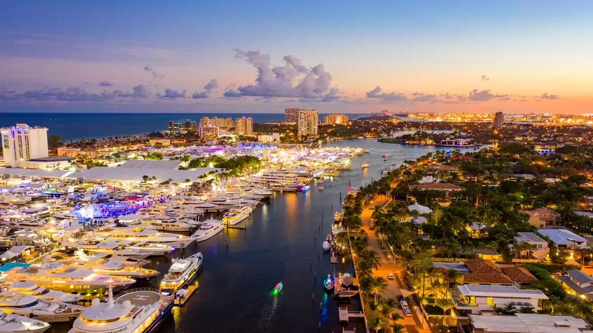 Aerial view of a harbor at sunset, with numerous illuminated yachts and a city in the background.