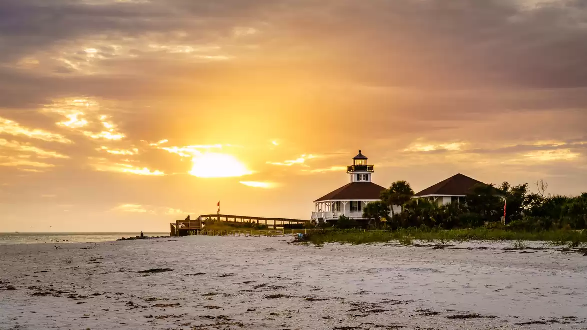 A picturesque sunset over a sandy beach with a pier and a lighthouse in the background.