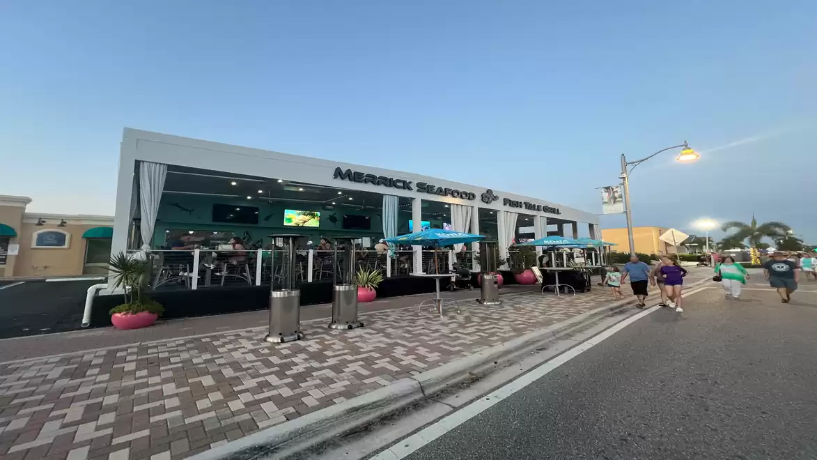 A modern restaurant with an outdoor terrace at sunset. People walk on a paved road in front of the building.