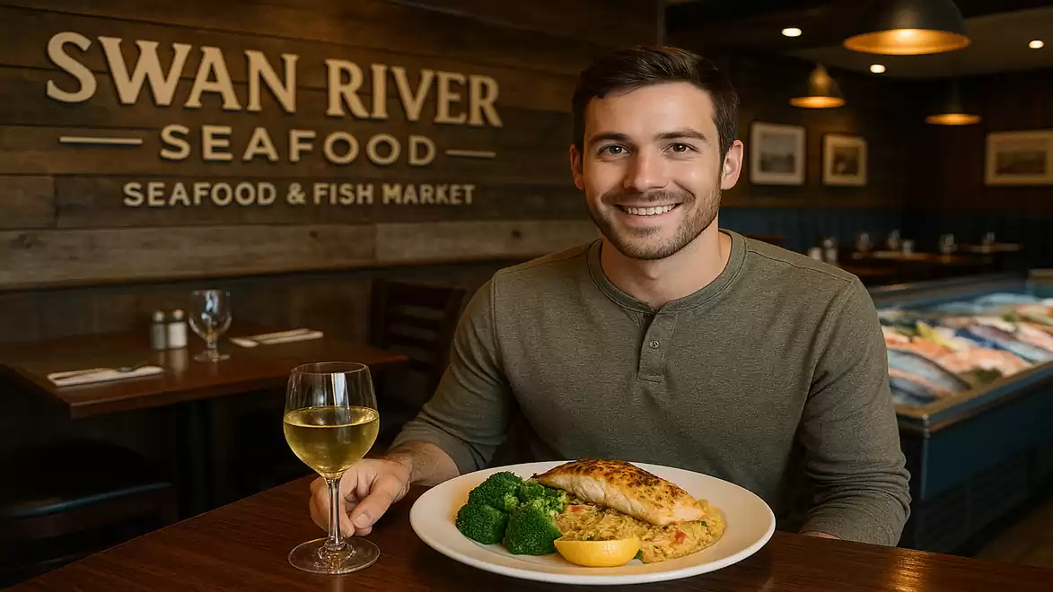 A smiling man sits in a restaurant called Swan River Seafood. He has a glass of white wine and a plate with grilled fish, broccoli, and lemon in front of him.