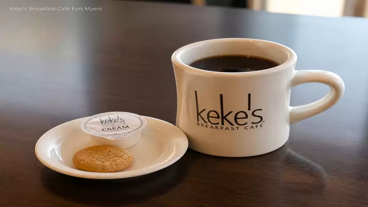 A white mug filled with black coffee on a table, next to a small plate with a cookie and a small plastic cup of cream. The mug and plate are labeled 'keke's Breakfast Cafe'.