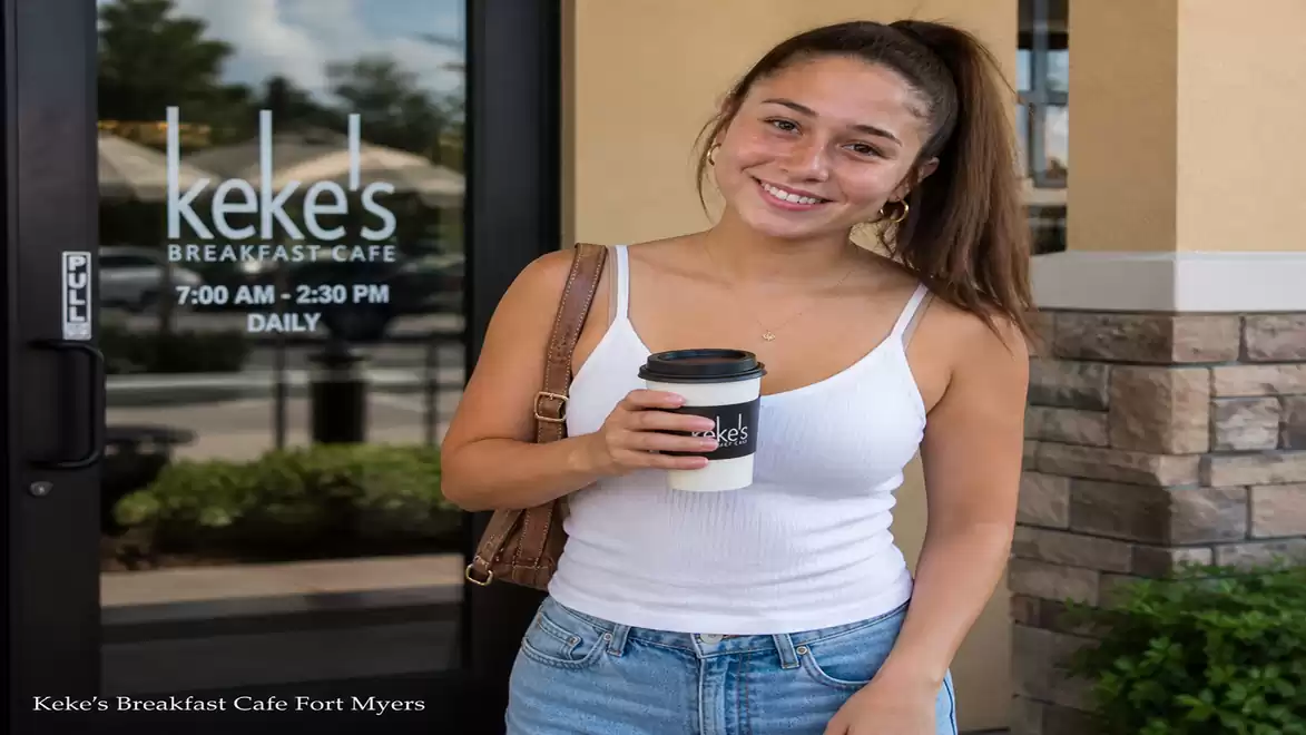 Woman in front of the entrance of Keke's Breakfast Café in Fort Myers, holding a coffee cup and smiling.
