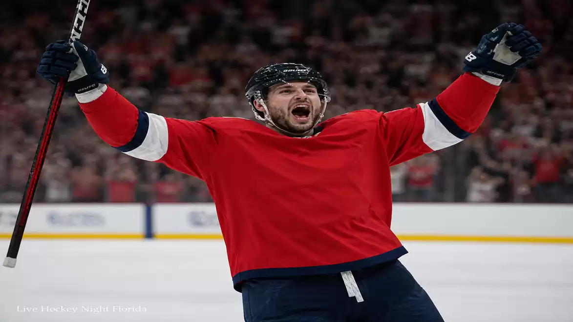 A hockey player in a red jersey celebrates with arms raised on the ice, with the crowd in the background cheering.