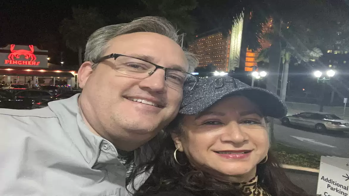 A smiling couple takes a selfie at night with illuminated buildings in the background.