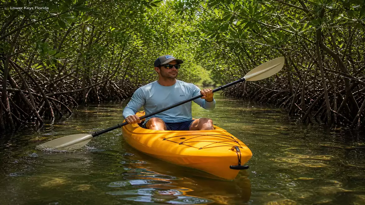 A man paddling an orange kayak through a dense mangrove forest with clear water, wearing sunglasses and a cap.