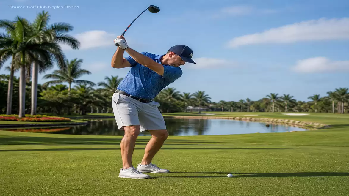 A golfer in blue shorts and a blue shirt swings his club on a well-manicured golf course with palm trees and a water hazard in the background.