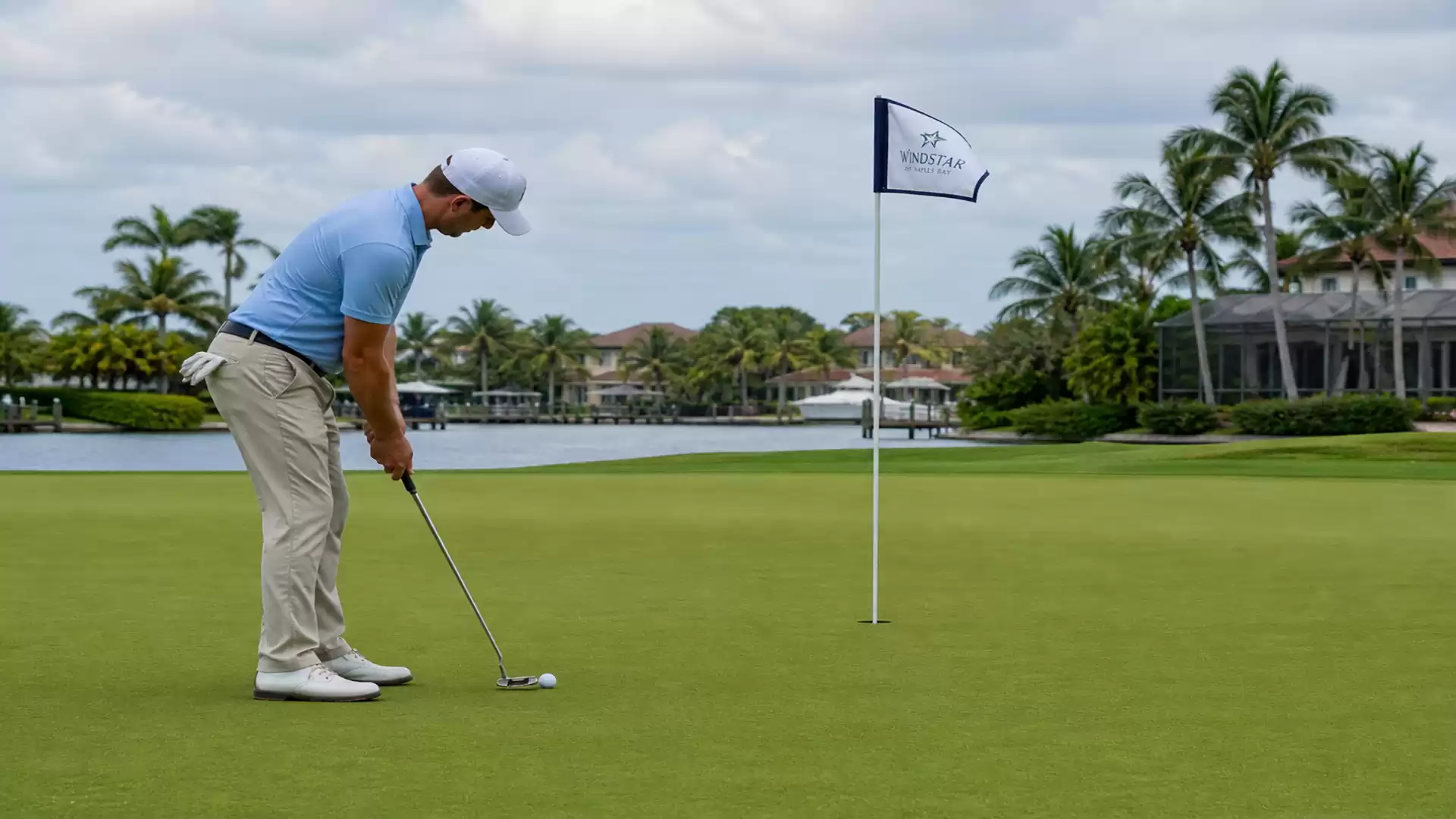 A golfer in beige pants and a blue shirt putts on a golf green with palm trees and a lake in the background.
