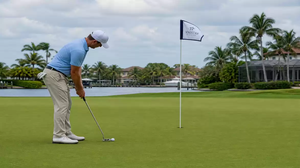 A golfer in beige pants and a blue shirt putts on a golf green with palm trees and a lake in the background.