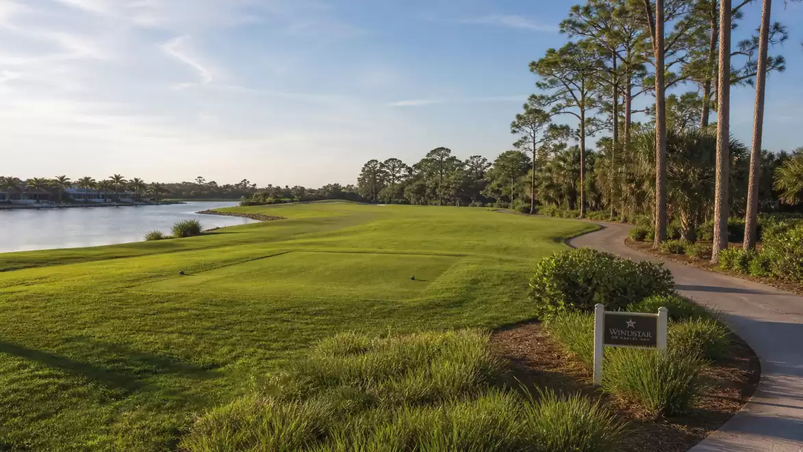 A picturesque golf course hole with a green fairway, trees, and a view of a lake under clear sky.