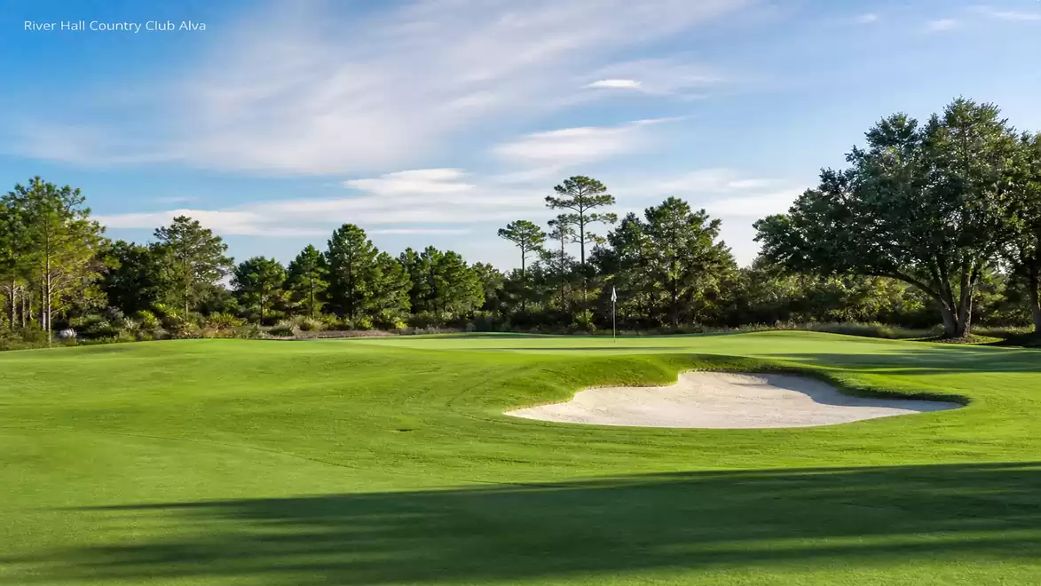 A well-maintained golf course with a sand bunker and green trees in the background under a blue sky.
