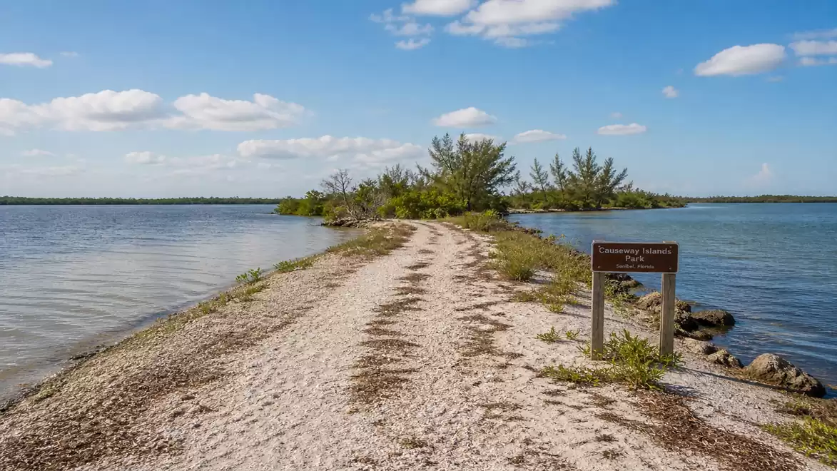 A narrow path leads over a sandbank to a small tree-covered island. Blue water extends on both sides of the path under a sky with a few clouds.